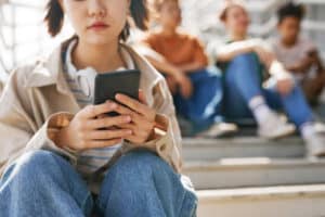 Closeup of teenage girl holding smartphone outdoors while sitting on metal stairs with group of friends